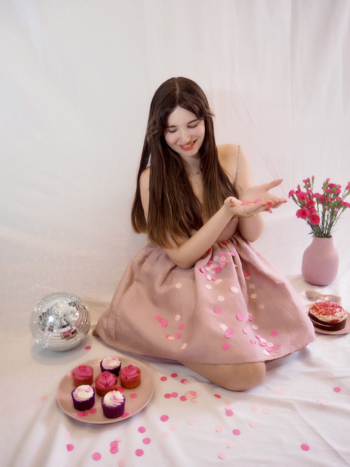 Girl wearing pink dress sits on the floor surrounded by confetti, pink cakes, pink flowers, and a disco ball. She's looking down at he left side at the pink cakes.