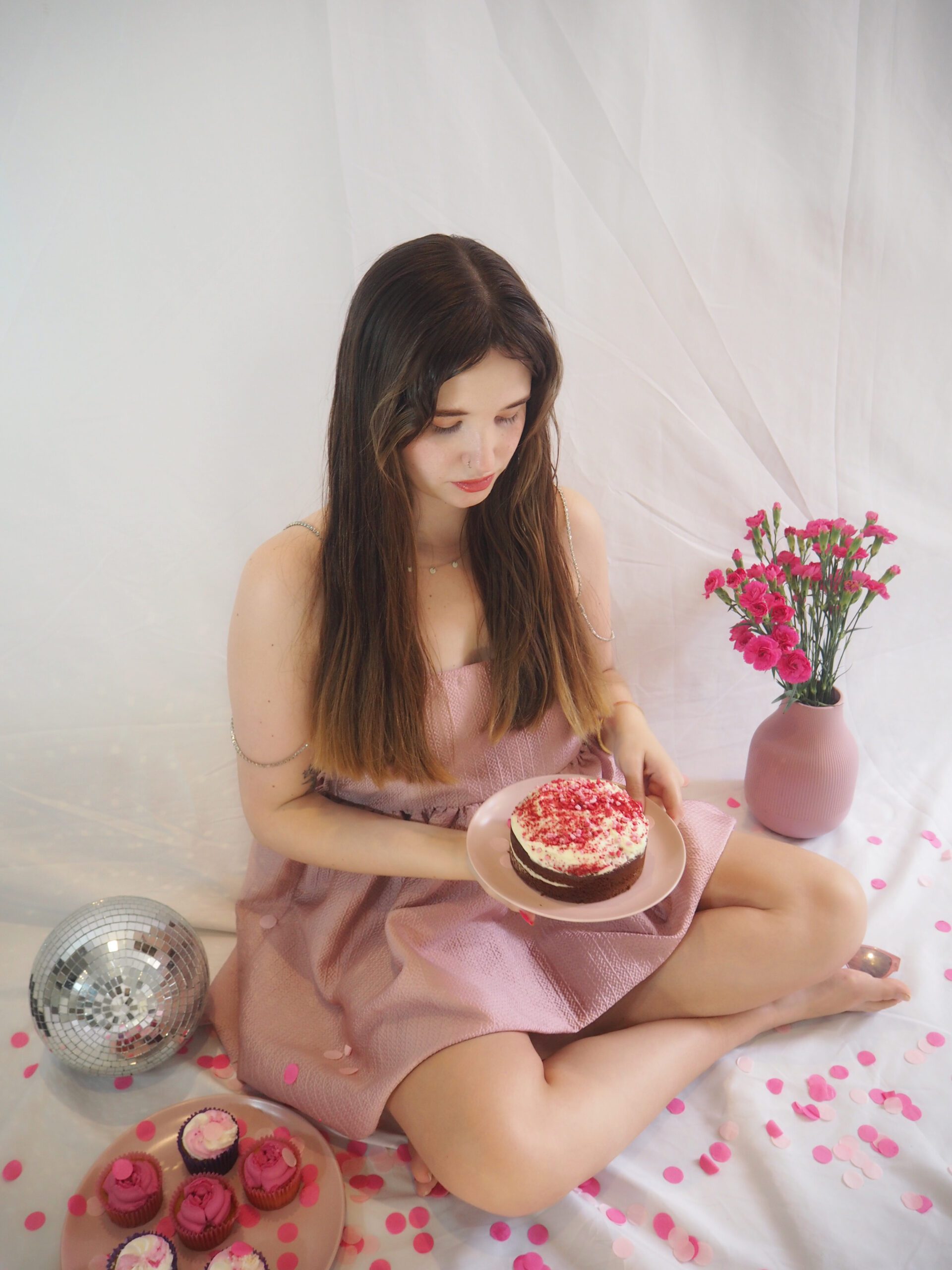 wedding business marketer and copywriter wearing a pretty pink party dress looking down at a pink cake in her hands. She's surrounded by wedding/party props including a disco ball, flowers, and confetti