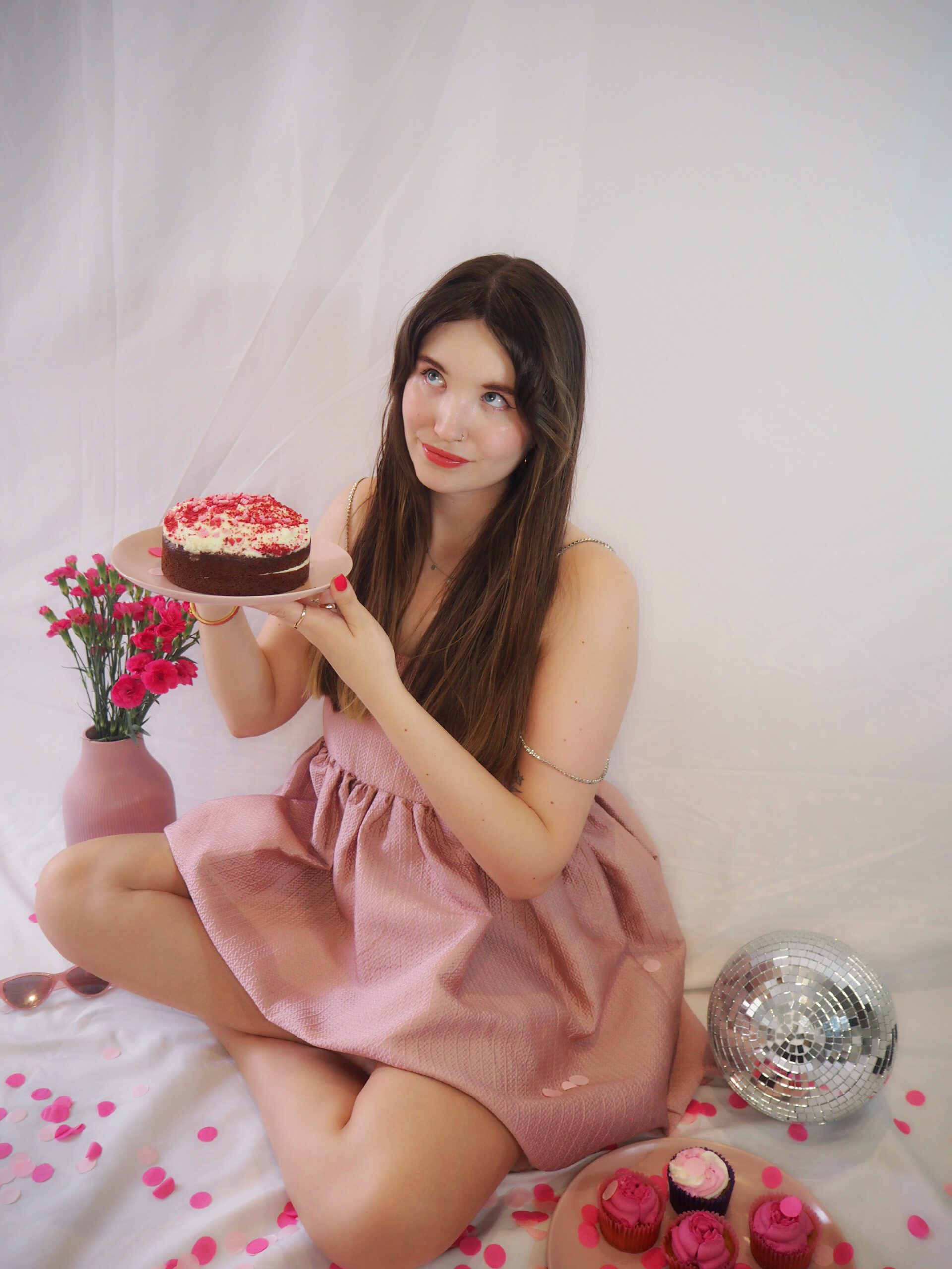Website copywriter wearing luxury pink party dress with silver straps. She's holding up a pink cake and is surrounded by confetti, cakes, and a disco ball. She's looking off to the left.