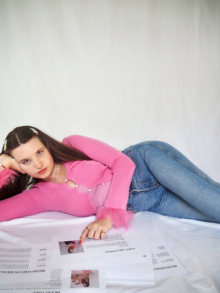 Girl wearing a pink top with fluffy sleeves and blue jeans looks at the camera as she lies down and points at brand strategy documents to start rebranding her copywriting business.