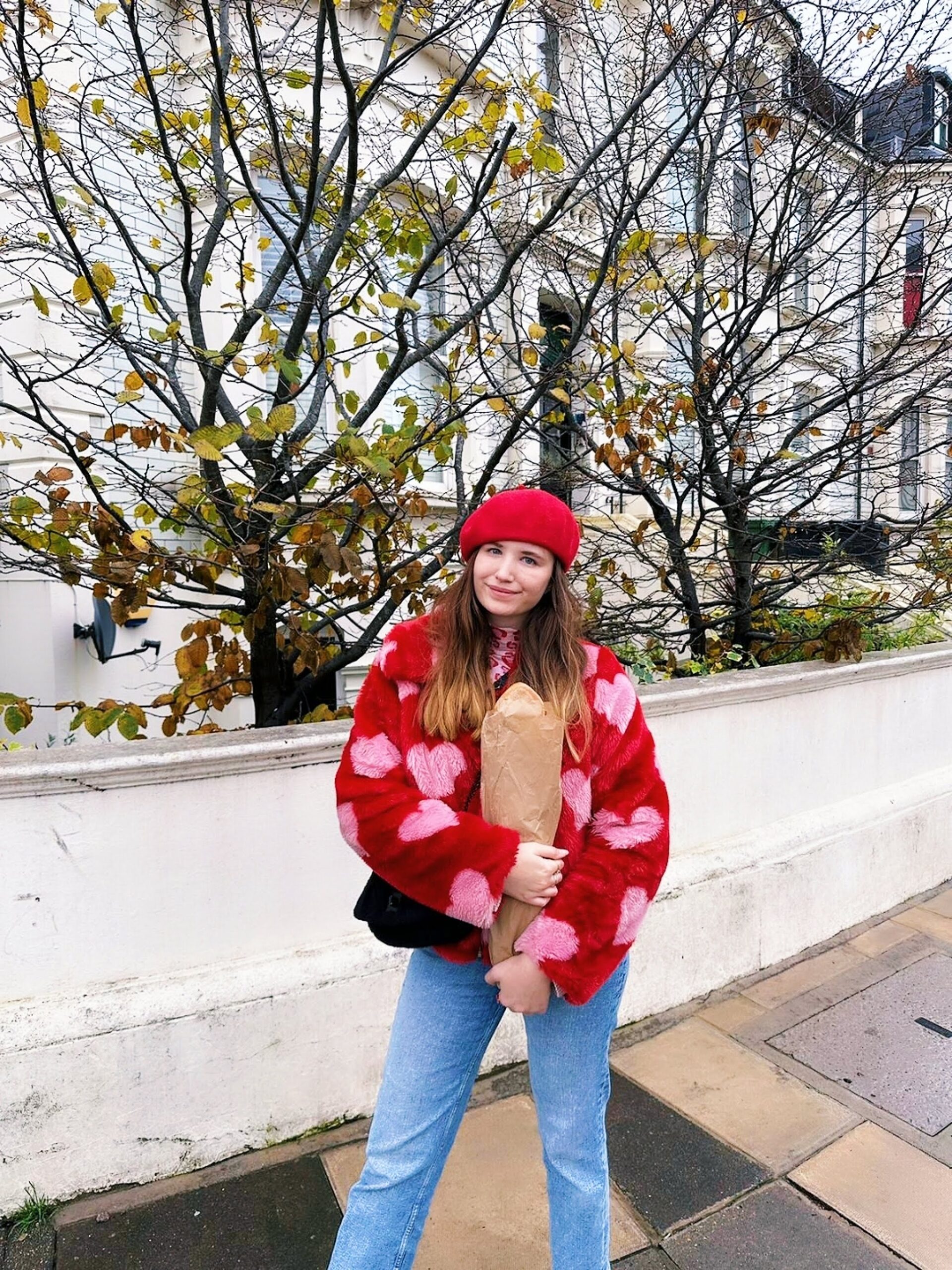 Website copywriter wearing a red beret and a heart print coat in red and pink and holding a loaf of bread while stood under trees.