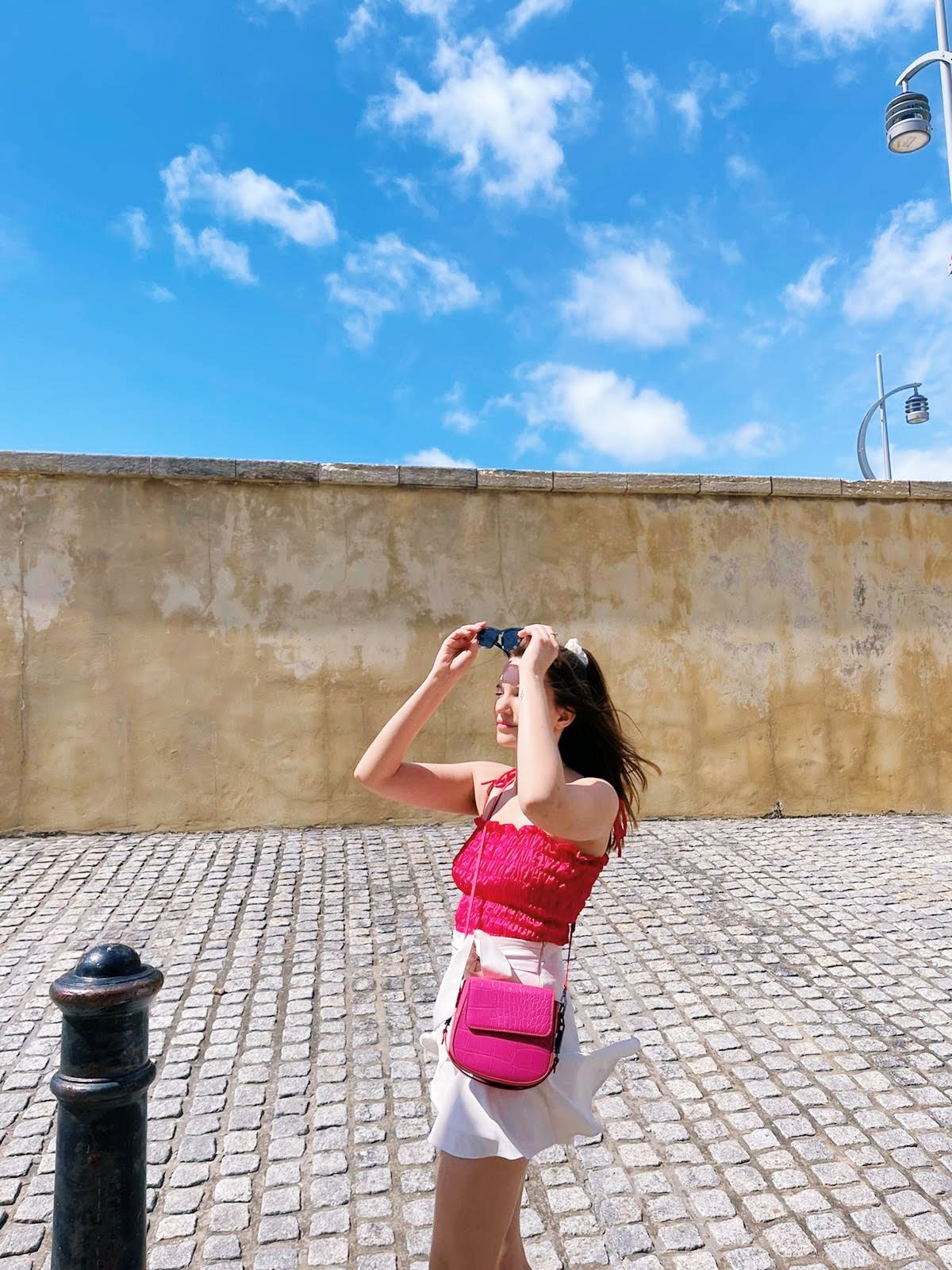 Female website content writer stood beneath a blue sky. She's wearing a pink top with a white skirt and has a pink bag across her body. She's holding her sunglasses up as she puts them on her head. There is a cobbled street behind her and a bollard to the left of the photo.