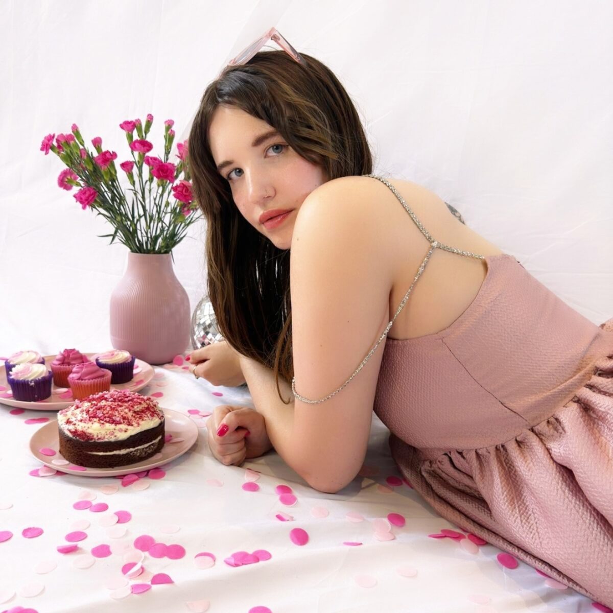 brand shoot for wedding industry copywriter. Brown haired girl wearing a pink dress leans across the floor with a white backdrop behind her and a vase of flowers and a wedding cake next to her on the left.