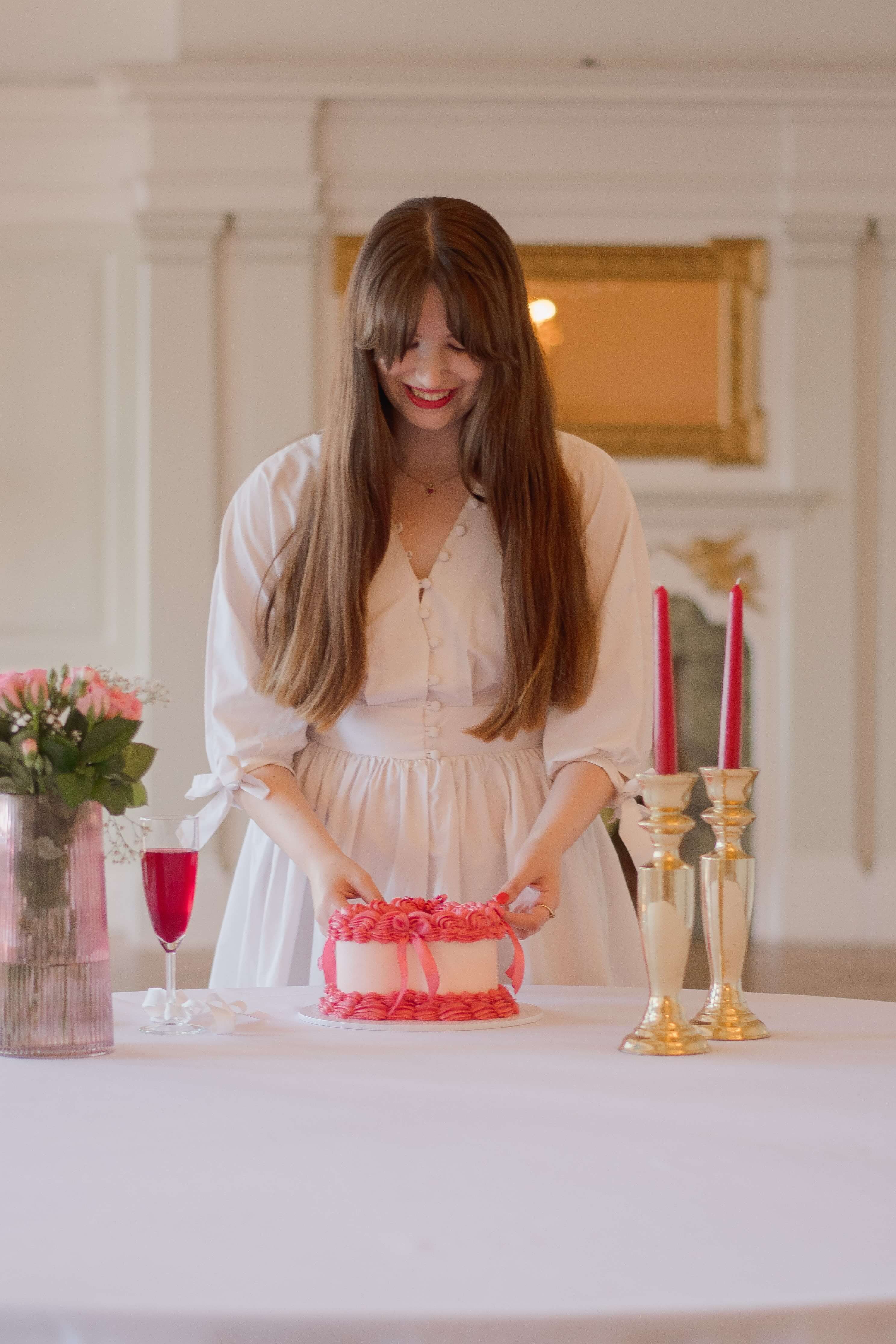 Wedding business brand photo of a girl standing behind a white table and table cloth. She's wearing a white dress with long sleeves and has long brown hair and bangs. On the table is a vintage heart-shaped cake, a champagne flute with re liquid, a pink vase with pink flowers, and gold candle sticks with red long candles in.