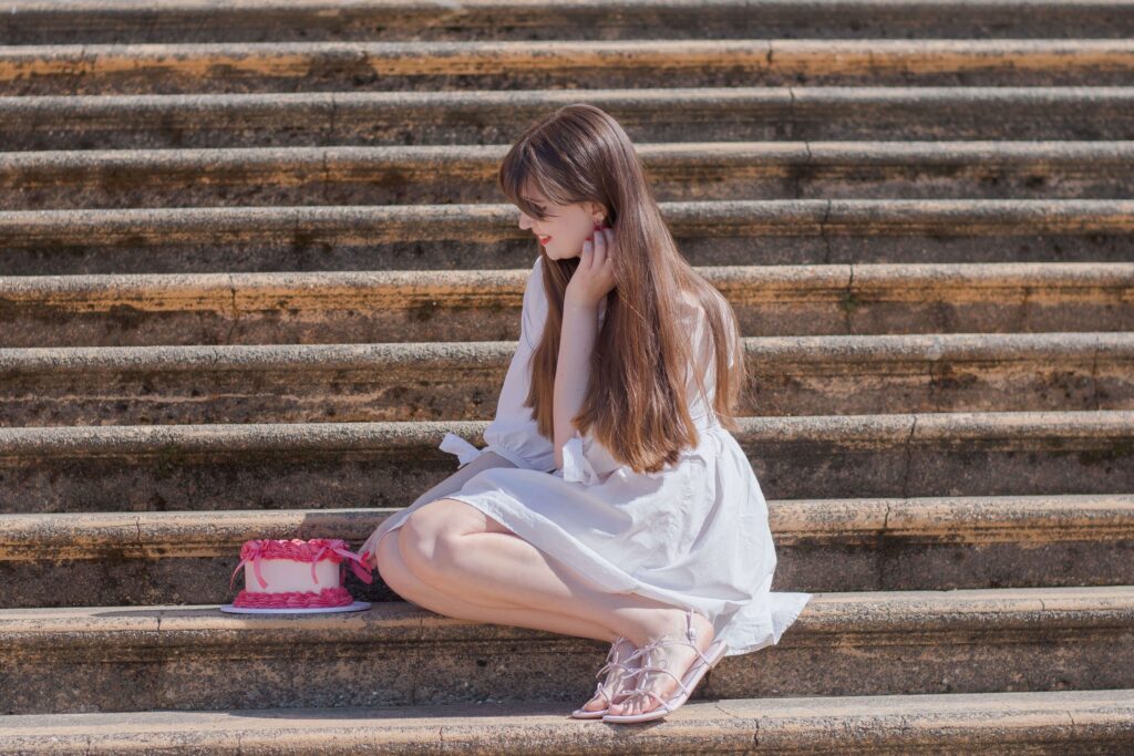Girl wearing a white wedding-inspired dress leans over a pink heart shaped cake while sat on the stairs of a wedding venue. She's wearing baby pink sandals with bows on.