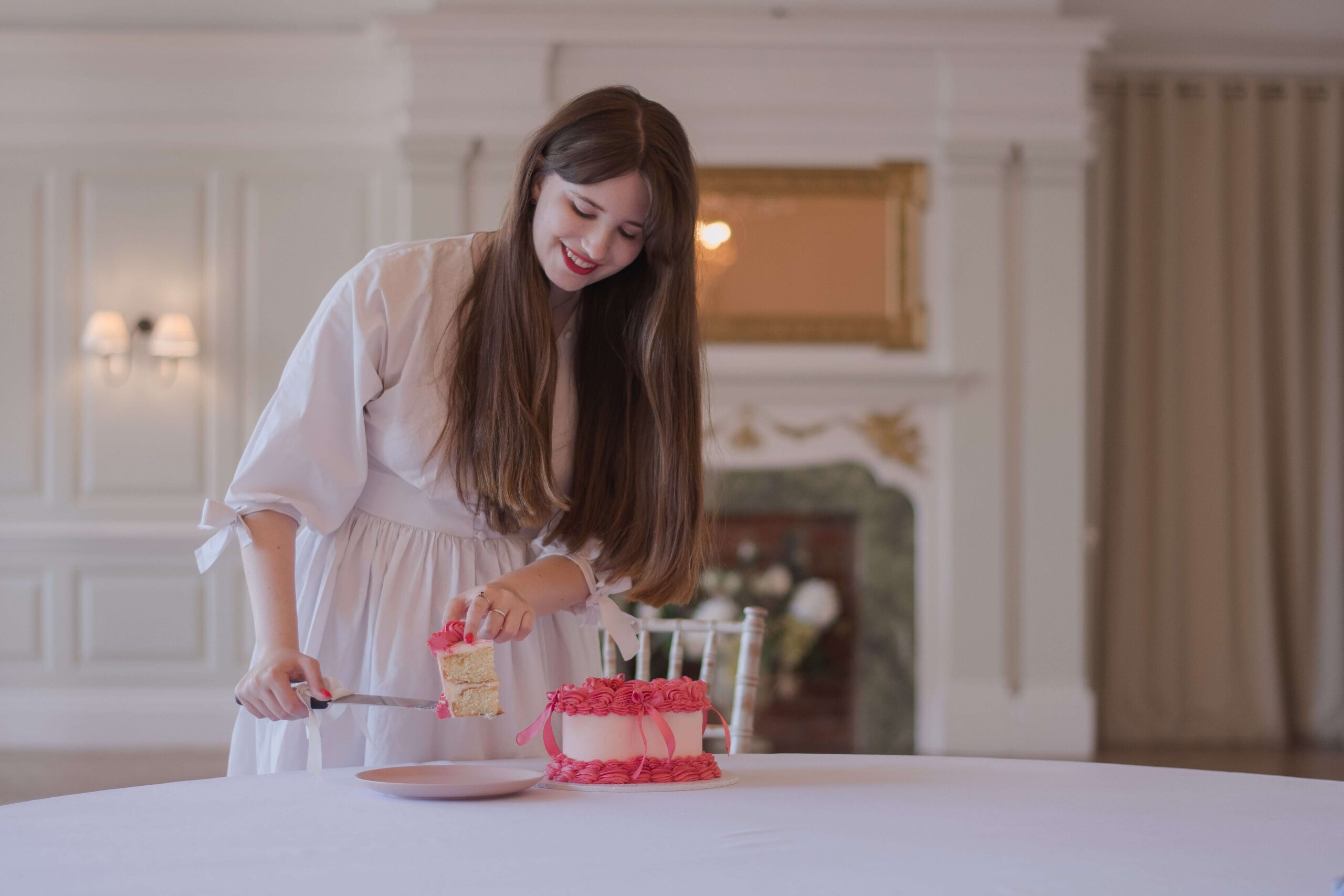 Wedding brand photo shoot of girl wearing a white dress and cutting a pink heart shaped wedding cake. She has long brown hair and bangs, and is stood behind a white table and in front of a fire place in a wedding venue.
