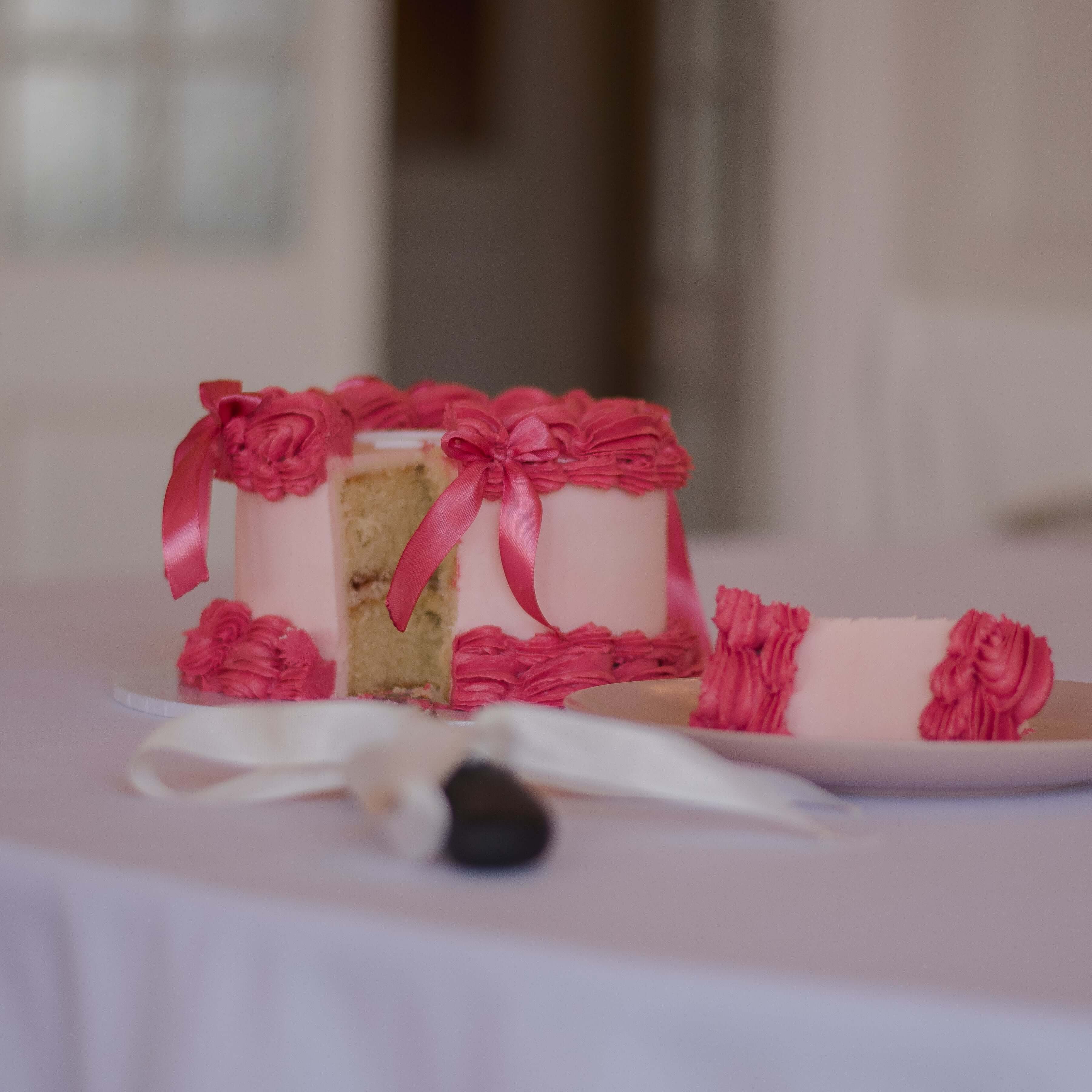 Pink and red heart-shaped cake with vintage-inspired frosting on a white table cloth with a black cake knife with a white ribbon tied around the handle.