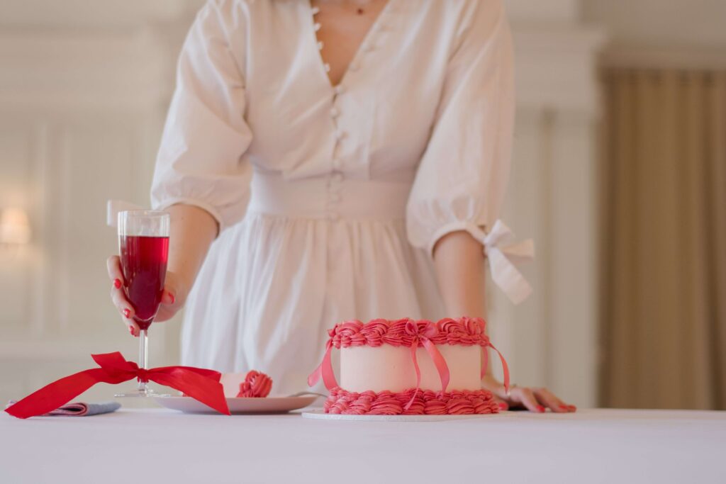 Wedding agency business photo shoot. A girl stands behind a white table with a pink, hear-shaped cake on it and a champagne flute with red liquid in and a red ribbon on. You can only see her torso and arms as she reaches out and holds the champagne flute.